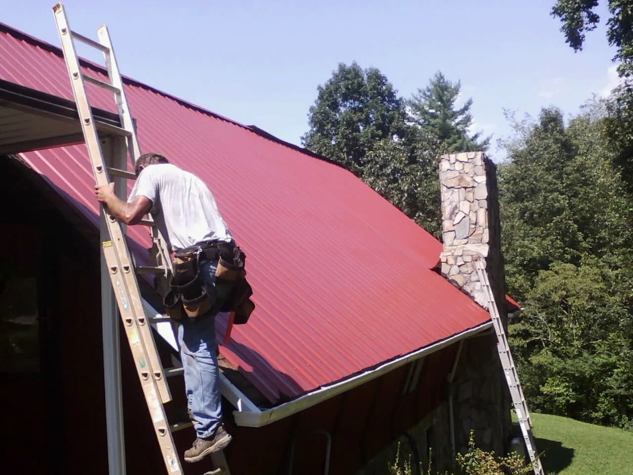 Roofer on a ladder beside a red metal roof and stone chimney — Soffit & Fascia Repair by Roofing Brothers in South Thomaston